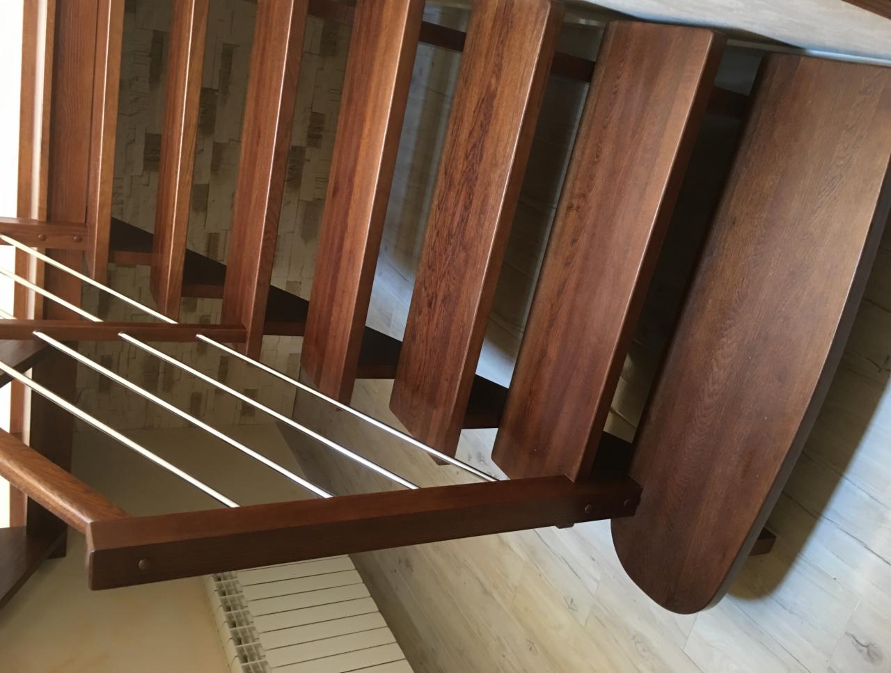 Wnętrze z drewnianymi schodami o ciepłym odcieniu brązu, z metalowymi prętami zabezpieczającymi, widoczne z góry, z fragmentem grzejnika w tle.