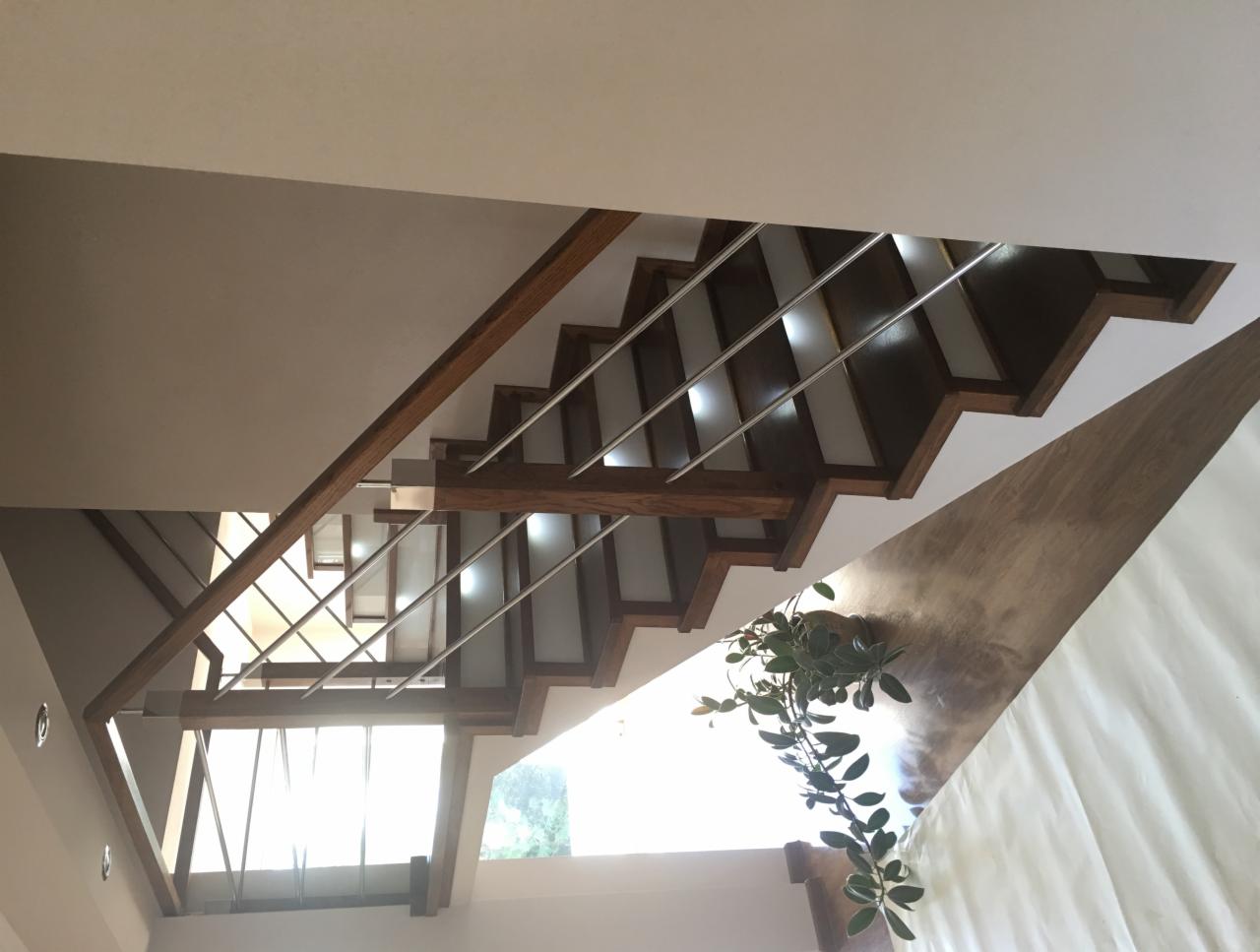 Wnętrze domu z nowoczesnymi schodami o drewnianych stopniach i metalowych barierkach, widok z dołu, z rośliną doniczkową na ścianie i oknem w tle.