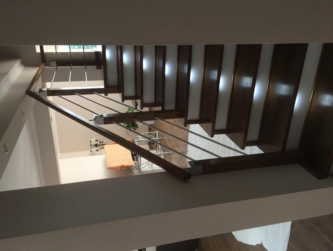 Wnętrze domu z widokiem na schody z drewnianymi stopniami i metalowymi poręczami, oświetlone światłem wpadającym przez okno na górze.