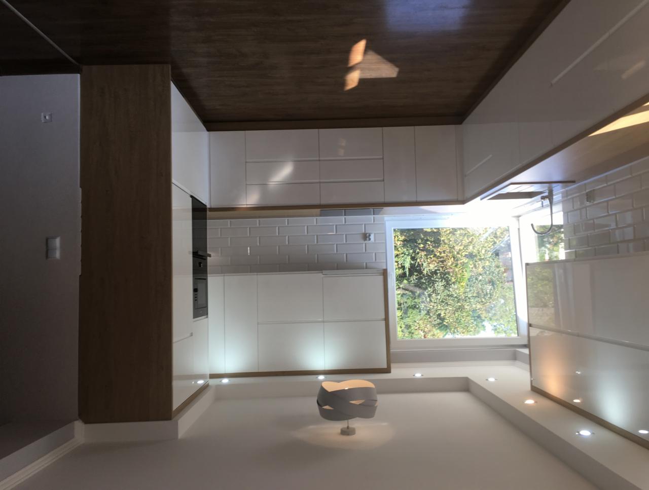 Nowoczesna, minimalistyczna kuchnia z białymi, lakierowanymi frontami, drewnianym blatem sufitowym i oświetleniem punktowym pod szafkami, widok na zieleń za oknem.