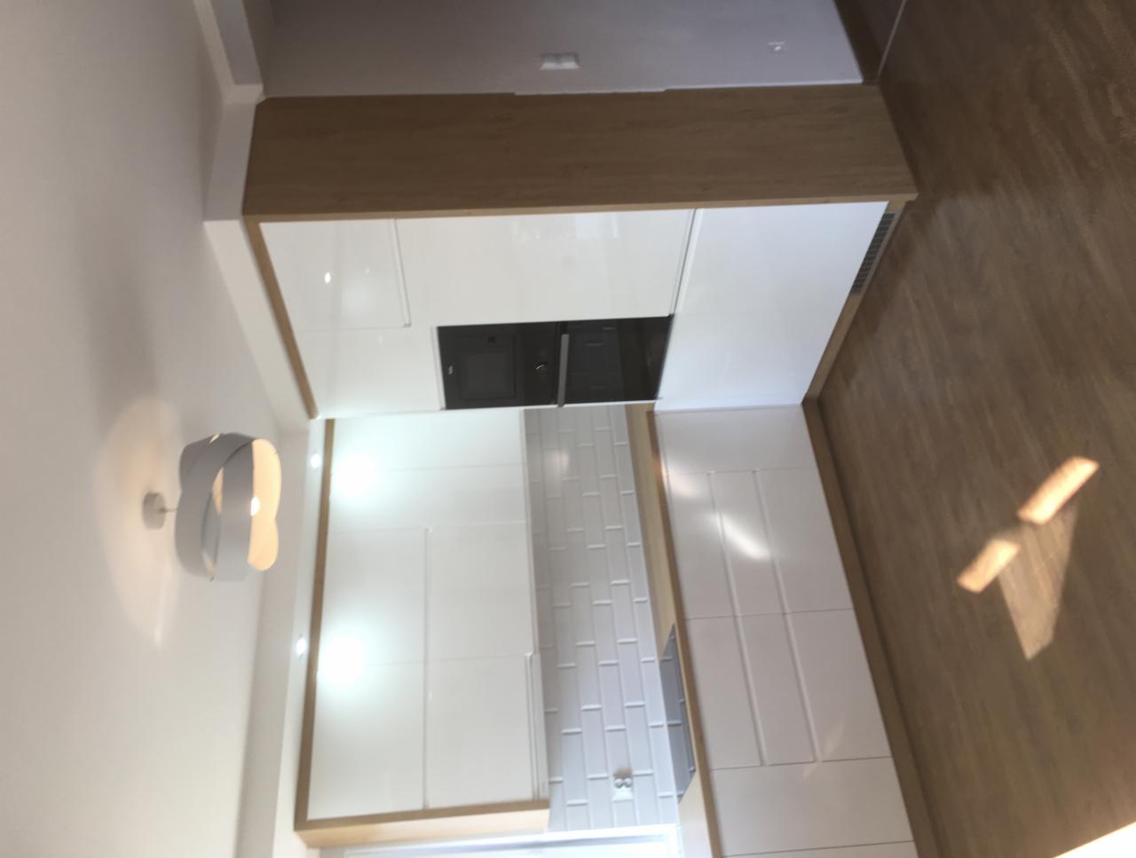 Nowoczesna kuchnia z białymi, błyszczącymi frontami szafek i drewnianym blatem, oświetlona designerskim kinkietem na ścianie, z widoczną zabudowaną płytą grzewczą i białymi płytkami na ścianie.