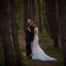 Fotografia Zaczarowane Kadry - Para młoda w lesie podczas sesji zdjęciowej, panna młoda opiera się o pana młodego, oboje ubrani w stroje ślubne, leśne otoczenie z promieniami słońca.