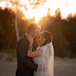 Fotografia Zaczarowane Kadry - Para młoda w objęciach, pocałunek na tle zachodzącego słońca, ona w koronkowej sukni i welonie, on w ciemnym garniturze, widoczny tatuaż na jej ramieniu i biżuteria.