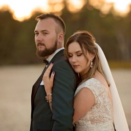 Fotografia Zaczarowane Kadry - Para młoda: panna młoda opiera głowę na ramieniu pana młodego w ciemnozielonym smokingu, złote światło zachodzącego słońca w tle.