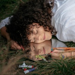 Fotografia Zaczarowane Kadry - Kobieta z kręconymi włosami leży na trawie obok lustra, w którym odbija się jej twarz malująca pędzlem na lustrze zieloną farbą; obok leżą tubki z farbami i pędzle.