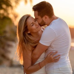 Fotografia Zaczarowane Kadry - Zakochana para w objęciach na plaży o zachodzie słońca, mężczyzna całuje kobietę w skroń, oboje ubrani na biało, widoczny podpis fotografa w lewym dolnym rogu.