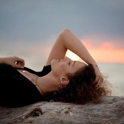 Fotografia Zaczarowane Kadry - Kobieta z kręconymi włosami w czarnej bluzce leży na spróchniałym pniu drzewa na tle zachodzącego słońca i pochmurnego nieba.