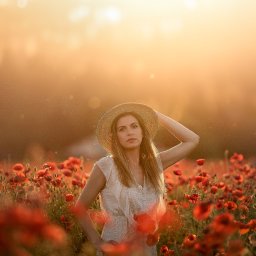 Fotografia Zaczarowane Kadry - Kobieta w słomkowym kapeluszu, stojąca na polu maków w ciepłym, zachodzącym słońcu, z ręką uniesioną do kapelusza i delikatnym uśmiechem.