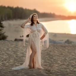 Fotografia Zaczarowane Kadry - Kobieta w koronkowej sukni i kapeluszu na plaży o zachodzie słońca, z widokiem na morze i las w tle.