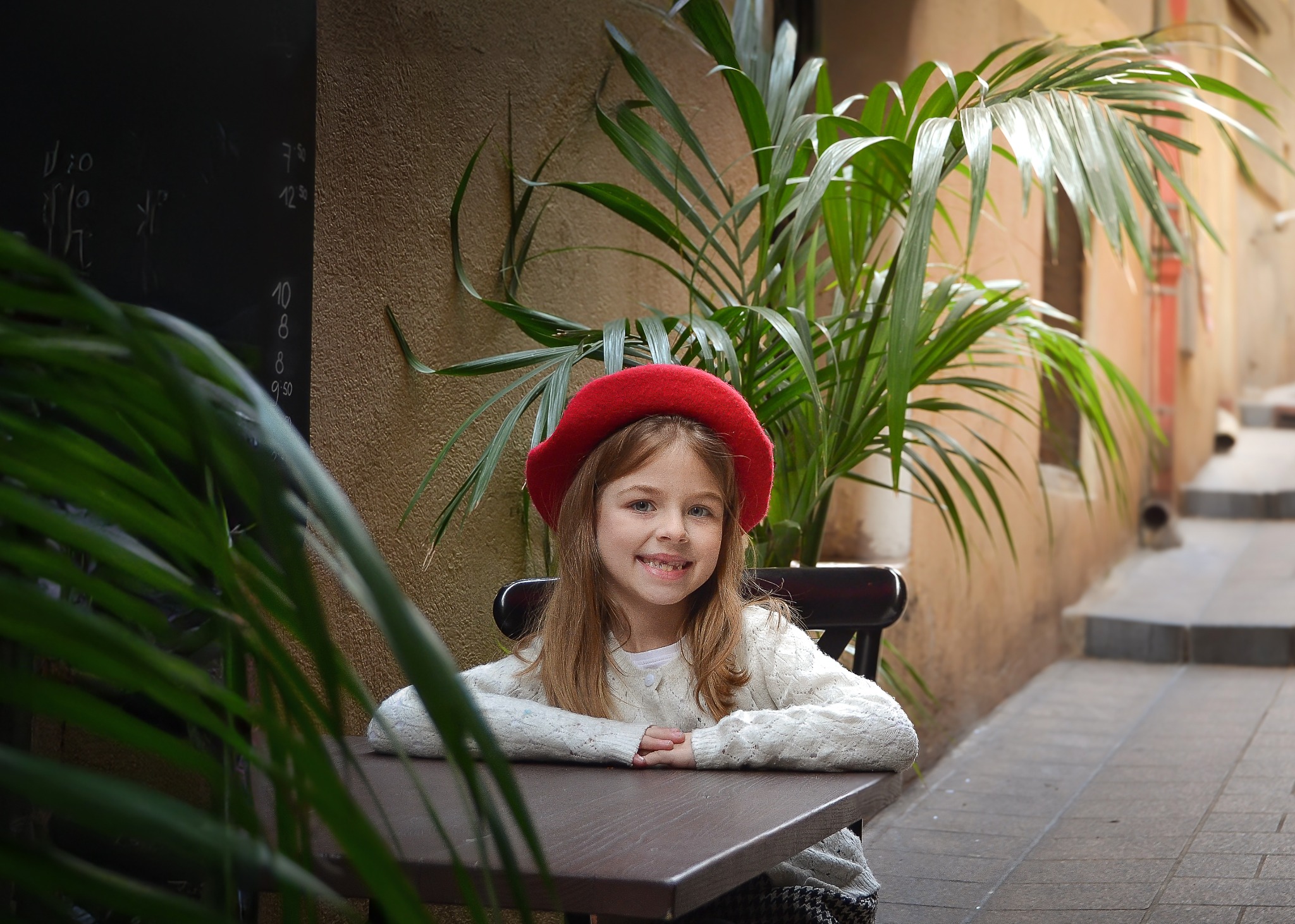 Uśmiechnięta dziewczynka w czerwonym berecie siedzi przy stoliku w kawiarni na tle ściany i rośliny doniczkowej.