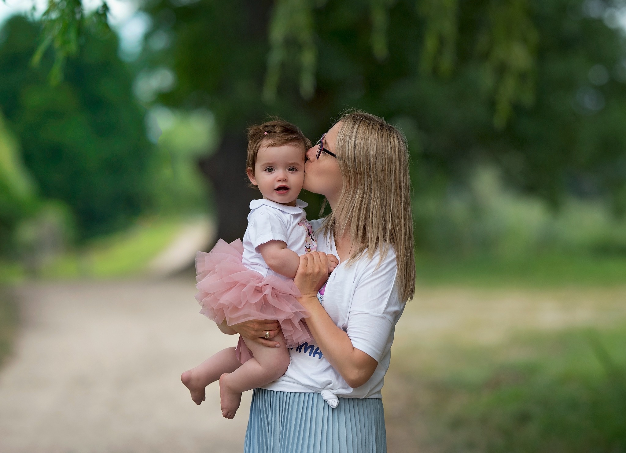 Mama w okularach całuje w policzek roczną córeczkę w tiulowej spódniczce podczas sesji w plenerze w Rotmance.