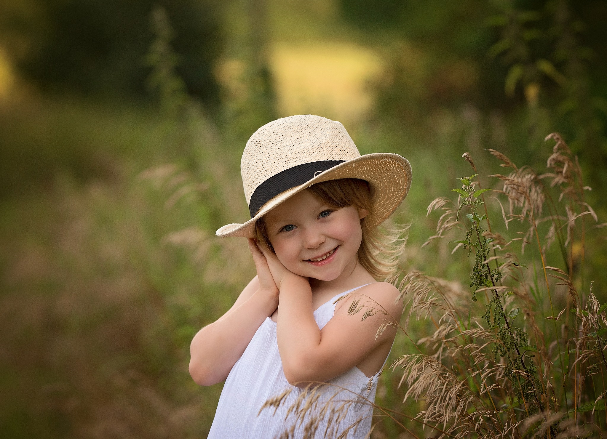 Uśmiechnięta dziewczynka w słomkowym kapeluszu, ubrana w białą sukienkę, pozuje w plenerze wśród traw i zieleni.