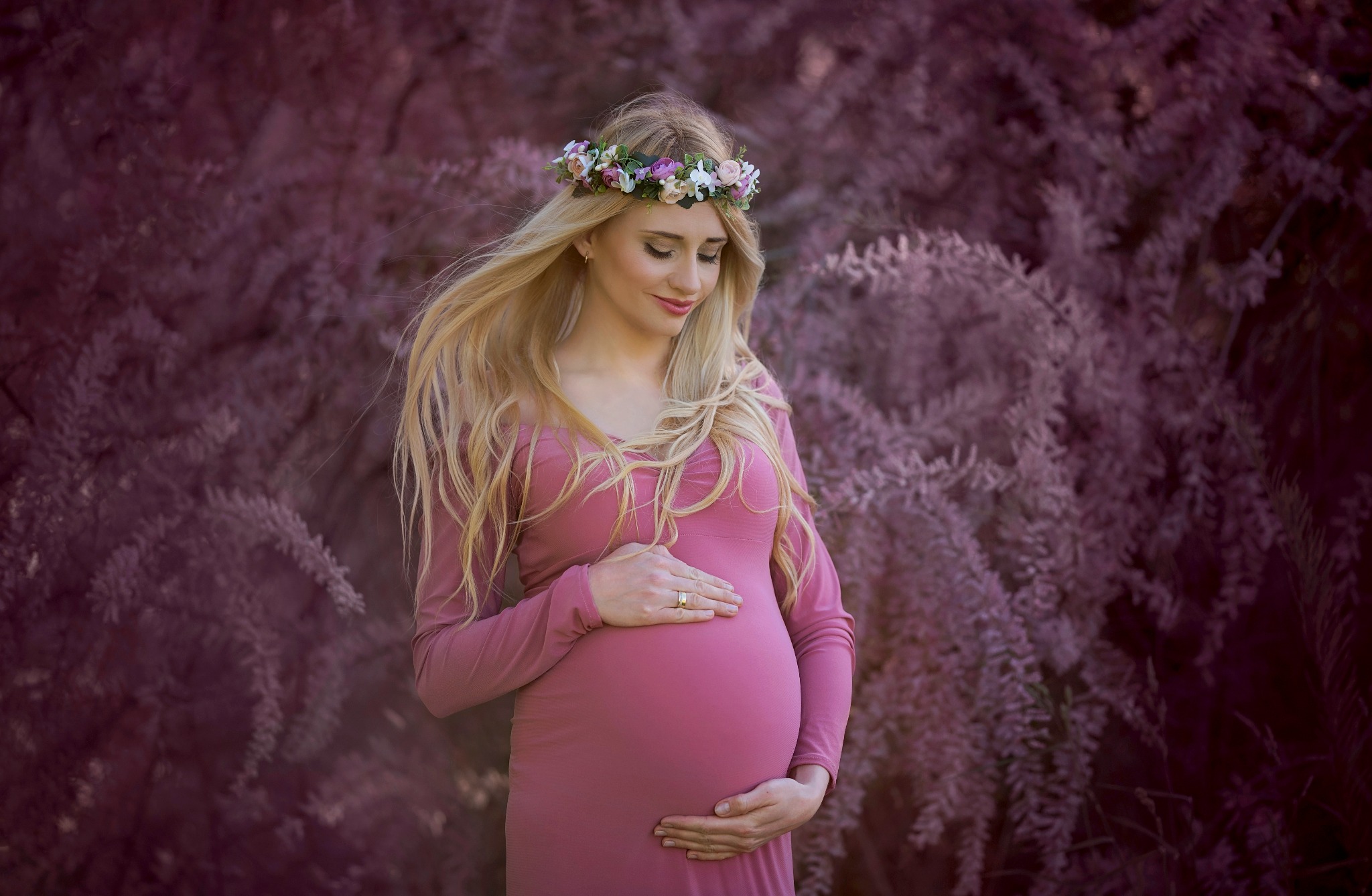 Kobieta w różowej sukience ciążowej, z wiankiem z kwiatów na głowie, delikatnie spogląda na swój brzuch na tle kwitnących, rozmytych krzewów.