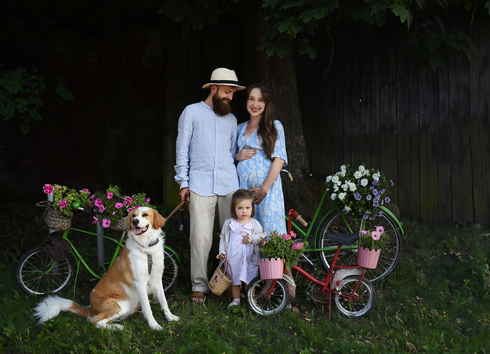 Uśmiechnięta para w ciąży z córeczką i psem pozuje przy rowerach ozdobionych kwiatami na tle drewnianej ściany.