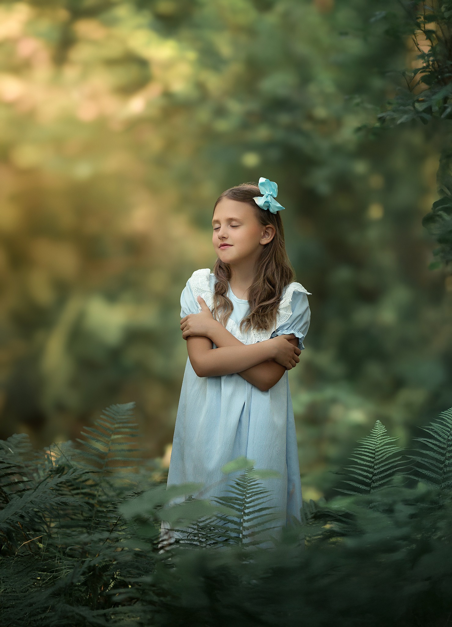 Dziewczynka w błękitnej sukience i z błękitną kokardą we włosach, stojąca pośród paproci w lesie, z zamkniętymi oczami i skrzyżowanymi ramionami.