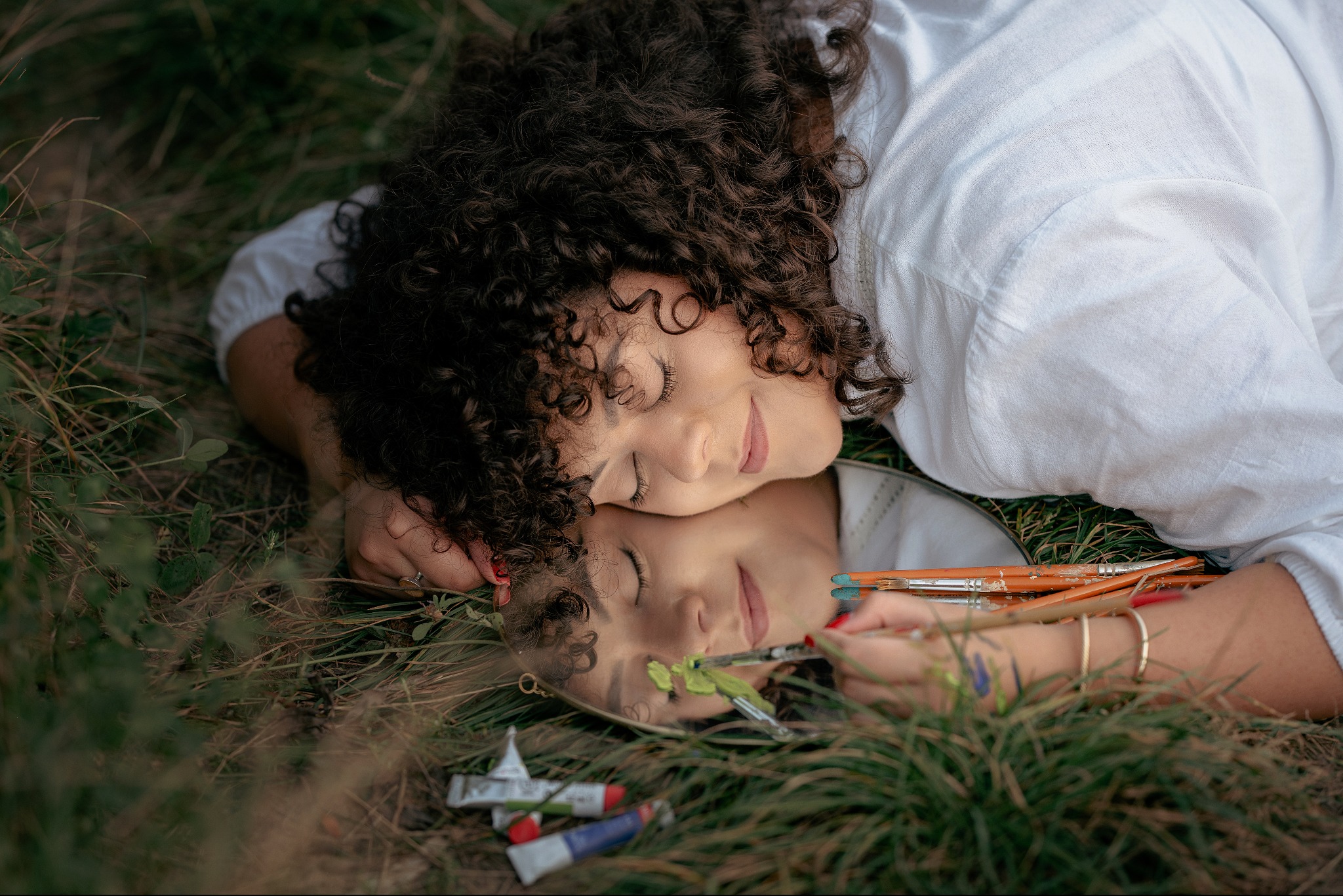 Kobieta z kręconymi włosami leży na trawie obok lustra, w którym odbija się jej twarz malująca pędzlem na lustrze zieloną farbą; obok leżą tubki z farbami i pędzle.