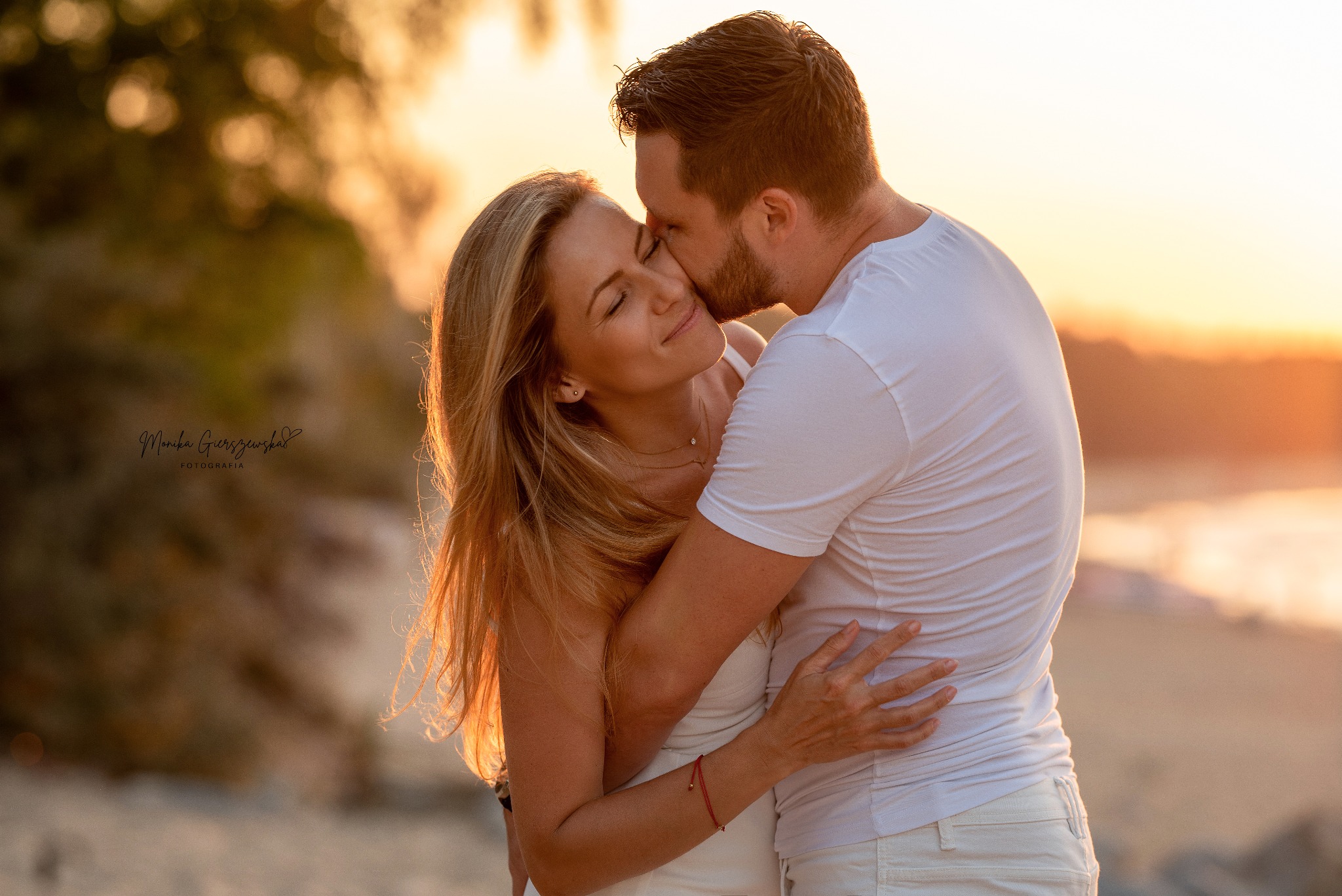 Zakochana para w objęciach na plaży o zachodzie słońca, mężczyzna całuje kobietę w skroń, oboje ubrani na biało, widoczny podpis fotografa w lewym dolnym rogu.