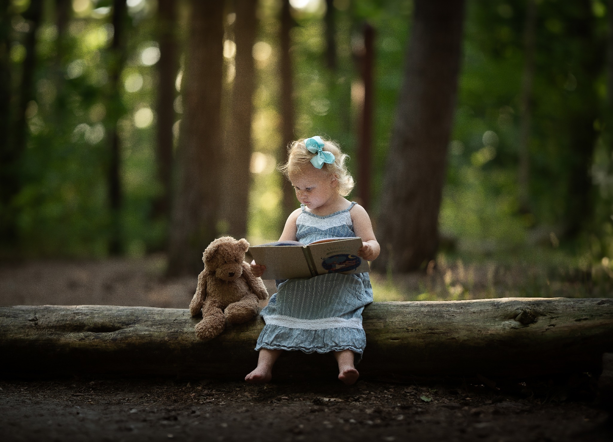 Dziewczynka z blond włosami i błękitną kokardą czyta książkę siedząc na powalonym pniu drzewa w lesie obok pluszowego misia.