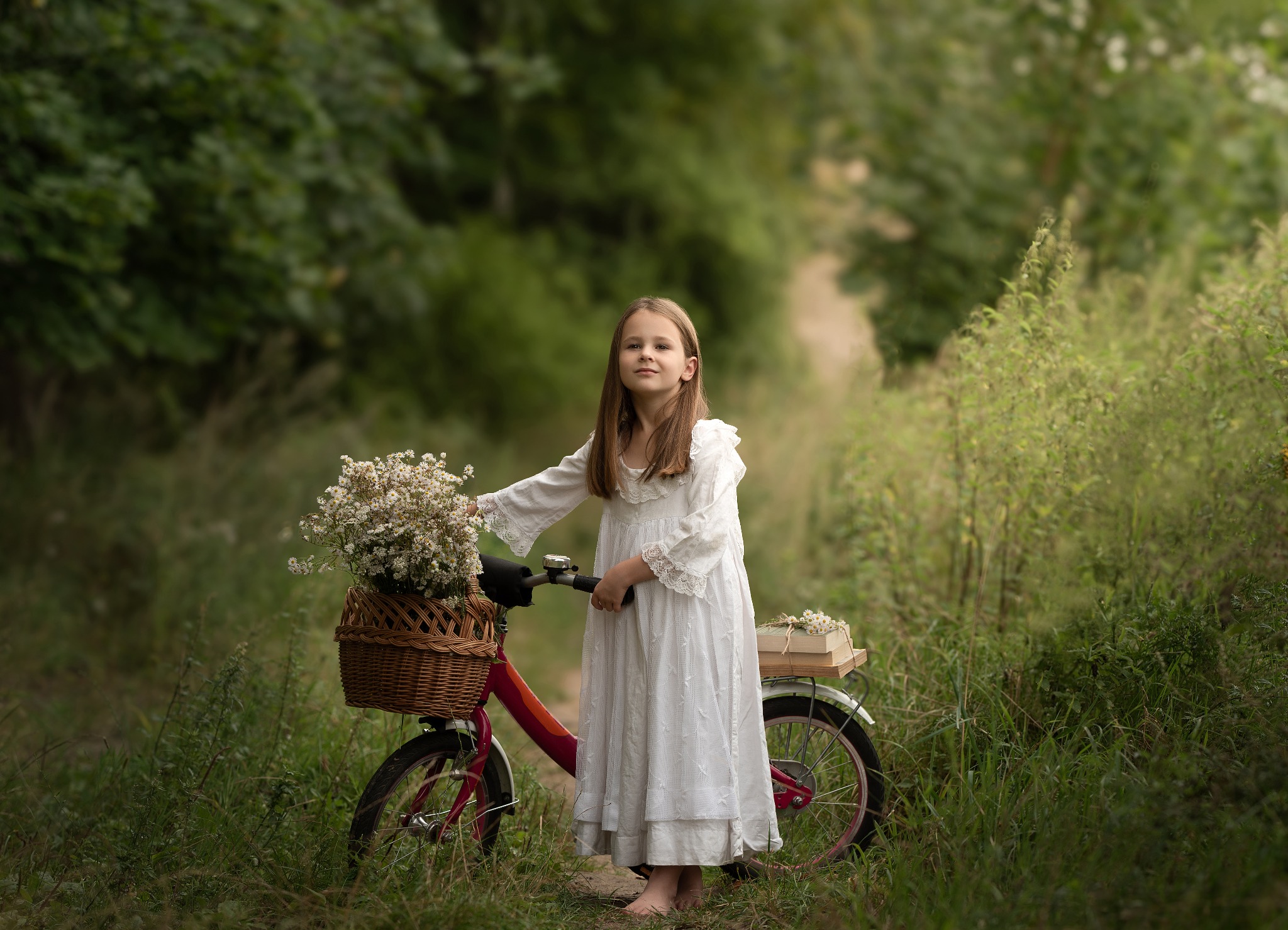 Dziewczynka w białej sukience stoi obok czerwonego roweru z koszykiem pełnym białych kwiatów na tle zieleni.