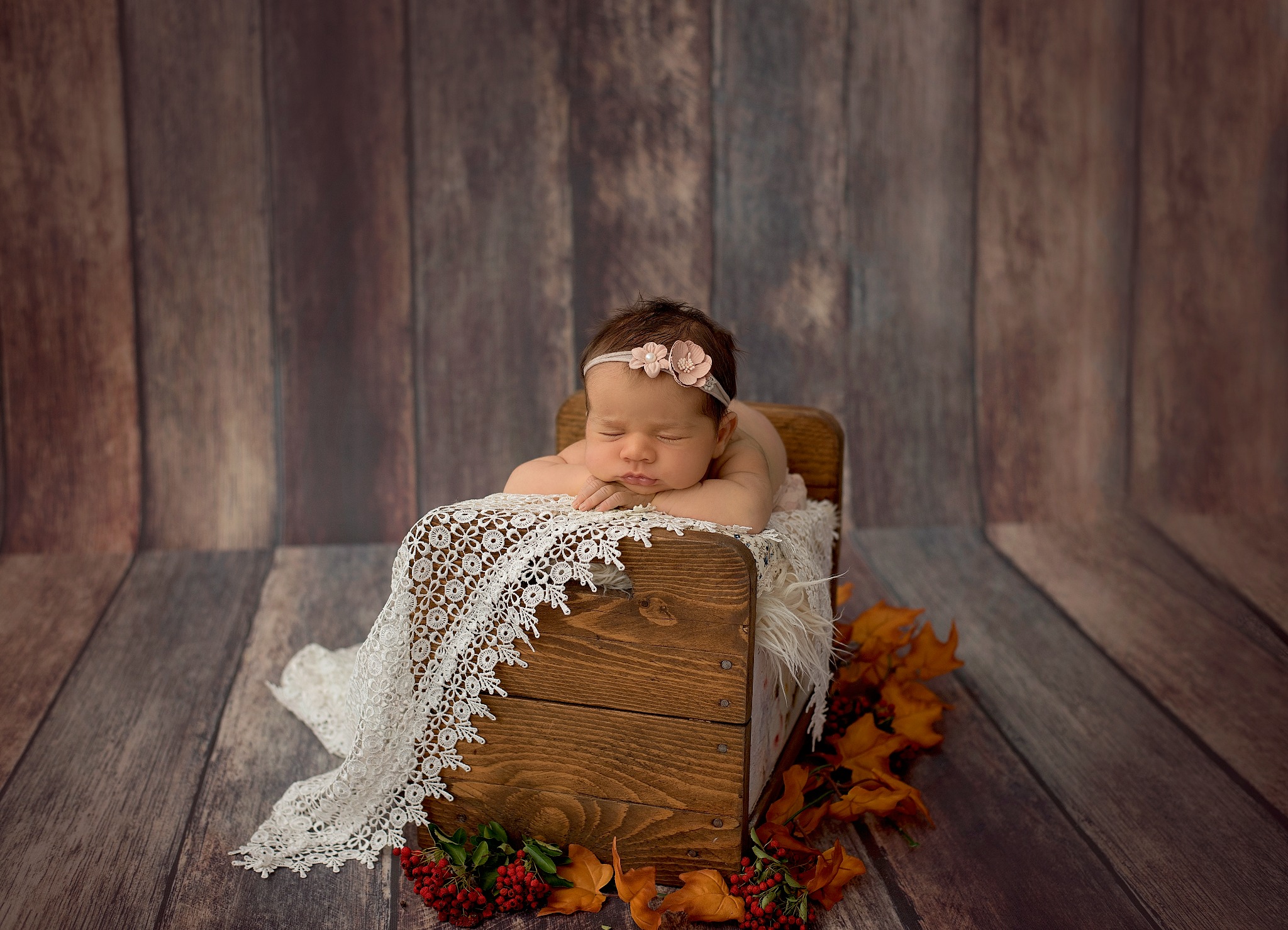 Śpiąca noworodka z opaską z kwiatkiem leży na małym drewnianym łóżeczku z koronkową narzutą, dekoracja z liści i jarzębiny, sesja w Rotmance, drewniane tło.