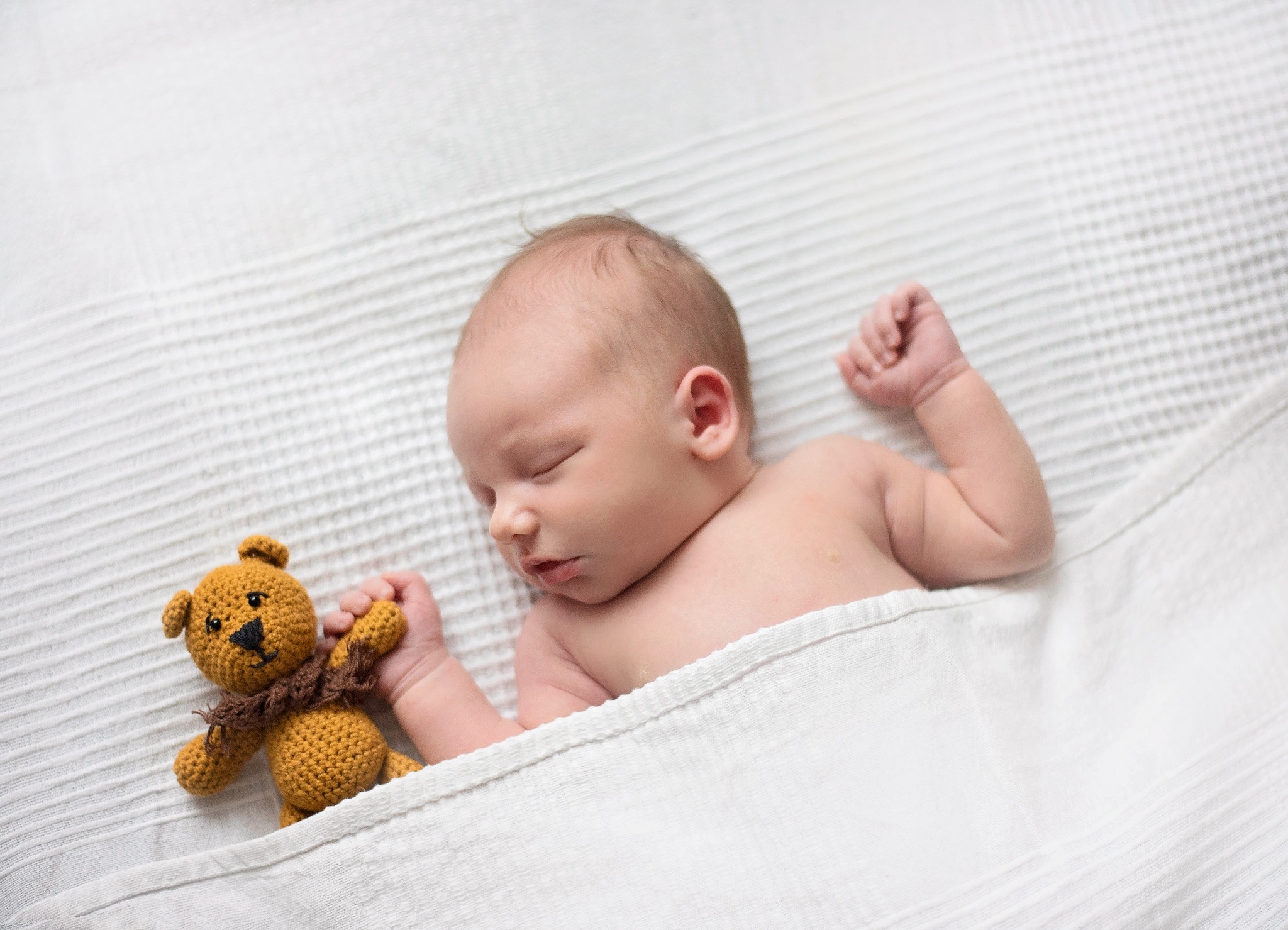 Śpiący noworodek owinięty w biały kocyk, trzymający małego, dzierganego, brązowego misia z szalikiem na białym tle.