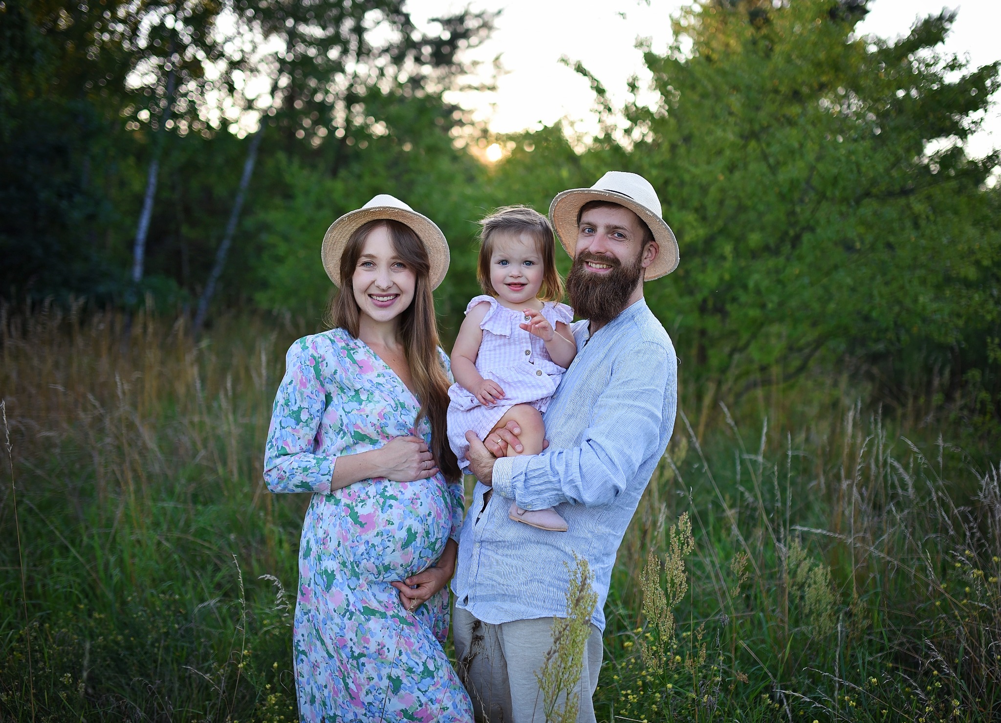 Uśmiechnięta, przyszła mama w kwiecistej sukience i słomkowym kapeluszu, stoi obok mężczyzny z brodą trzymającego na rękach małą dziewczynkę w różowej sukience, na tle zieleni.