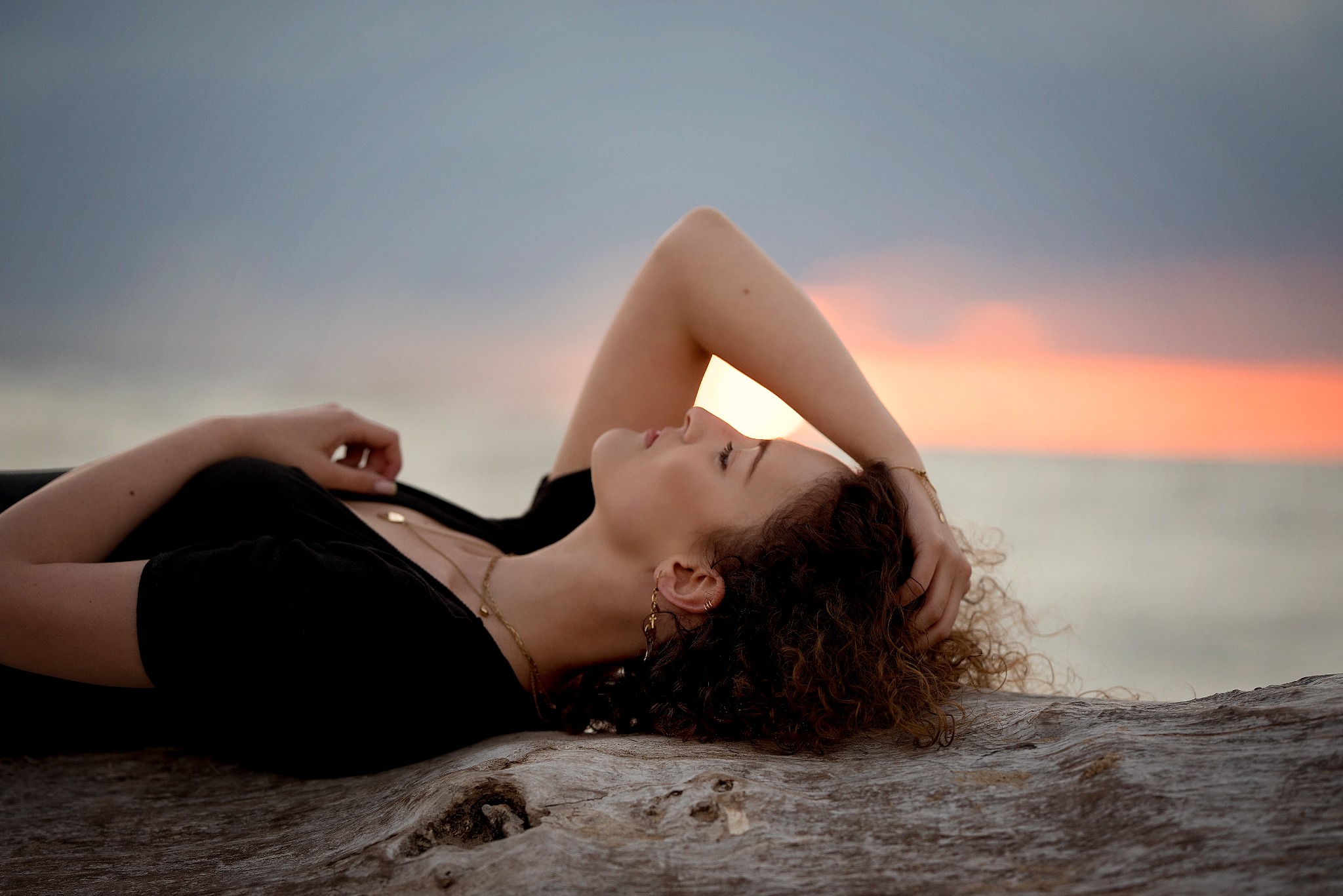 Kobieta z kręconymi włosami w czarnej bluzce leży na spróchniałym pniu drzewa na tle zachodzącego słońca i pochmurnego nieba.