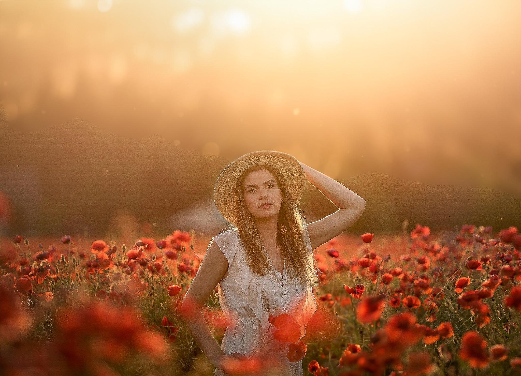 Kobieta w słomkowym kapeluszu, stojąca na polu maków w ciepłym, zachodzącym słońcu, z ręką uniesioną do kapelusza i delikatnym uśmiechem.