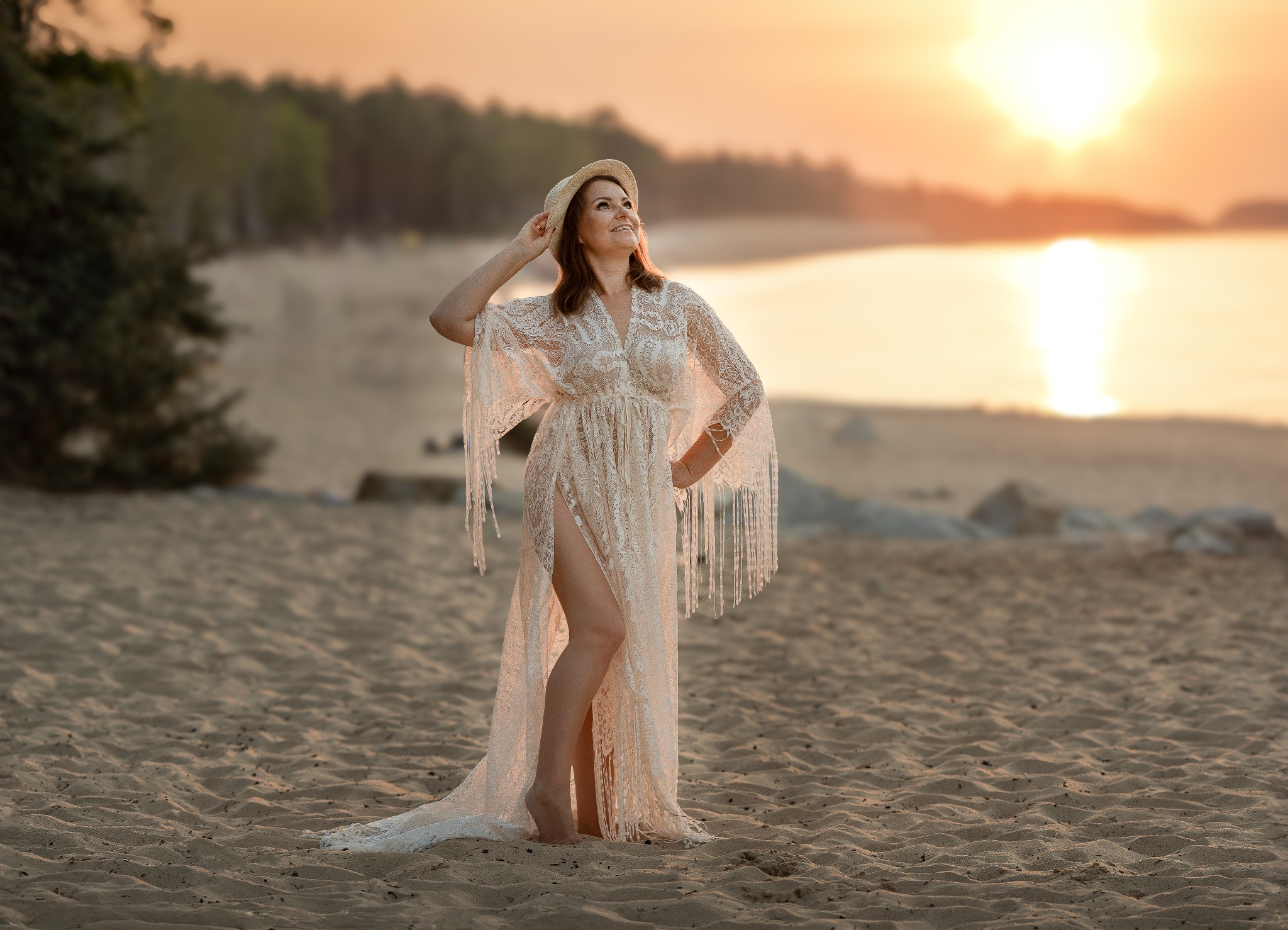 Kobieta w koronkowej sukni i kapeluszu na plaży o zachodzie słońca, z widokiem na morze i las w tle.