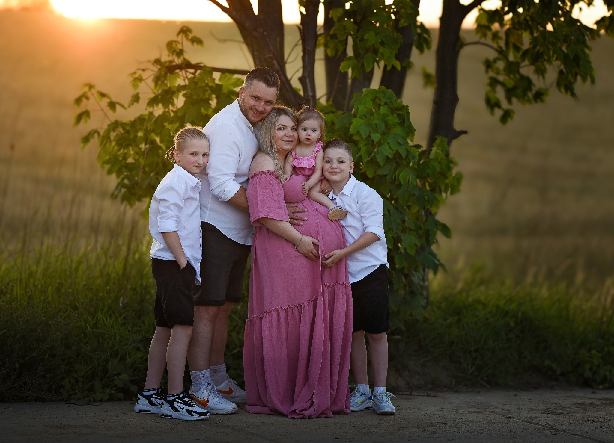 Rodzinne zdjęcie w plenerze przy zachodzącym słońcu: ciężarna kobieta w różowej sukience otoczona mężem i trójką dzieci, najmłodsze dziecko trzymane na rękach przez matkę.