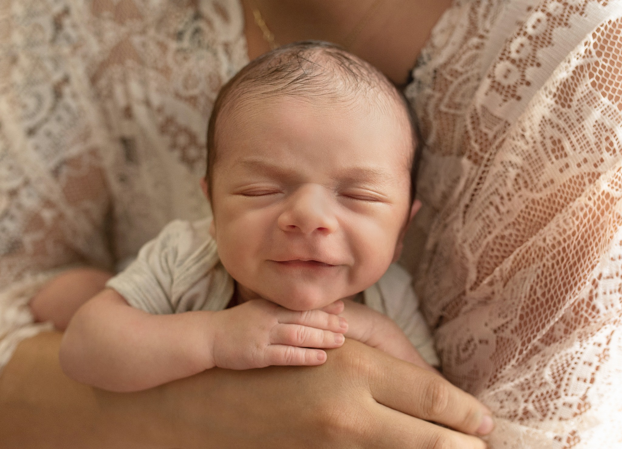 Noworodek z zamkniętymi oczami, delikatny uśmiech, trzymany w ramionach osoby ubranej w koronkową bluzkę, subtelne oświetlenie.
