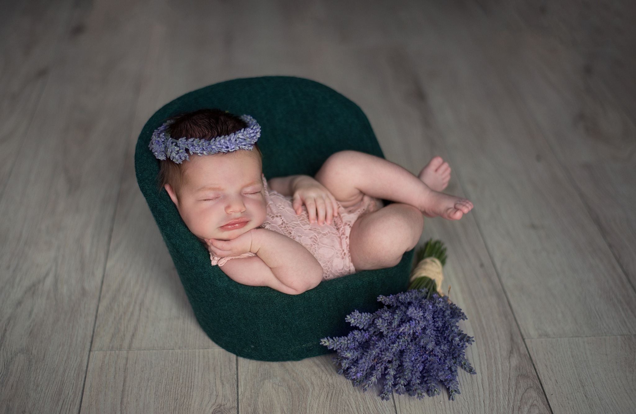 Śpiący noworodek w zielonym fotelu, ubrany w koronkowy kombinezon i wianek z lawendy, obok leży bukiet lawendy, całość na tle drewnianej podłogi.