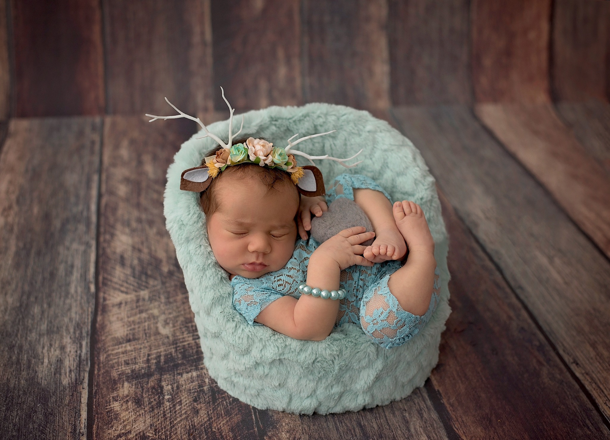 Śpiący noworodek w niebieskim koronkowym ubranku i opasce z rogami renifera, ułożony w miętowym pluszowym gnieździe na tle drewnianej podłogi.