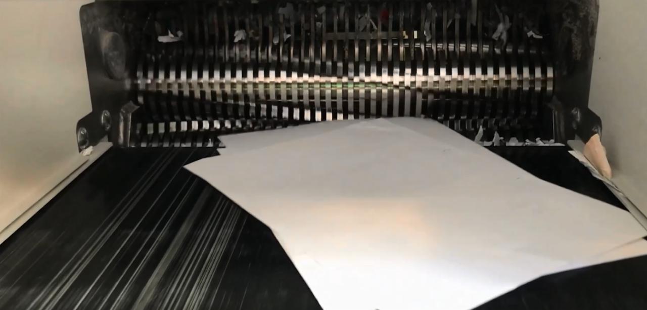 Białe kartki papieru w podajniku niszczarki przemysłowej, z widocznymi ostrzami tnącymi i strzępami dokumentów wewnątrz.