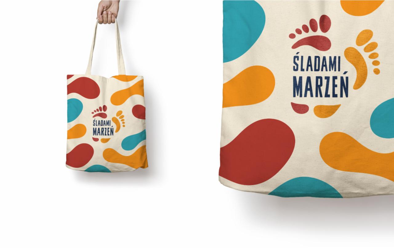 Beżowa torba bawełniana z nadrukiem logo festiwalu podróżniczego 'Śladami Marzeń', z kolorowymi plamami w kształcie stóp, trzymana w dłoni na białym tle.