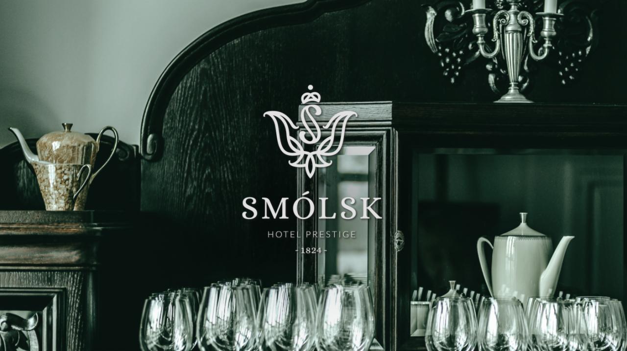 Elegancka aranżacja wnętrza hotelu Smólsk Prestige z ciemnym, drewnianym kredensem, kryształowymi kieliszkami, porcelanową zastawą i dekoracyjnym świecznikiem, podkreślona logo hotelu.