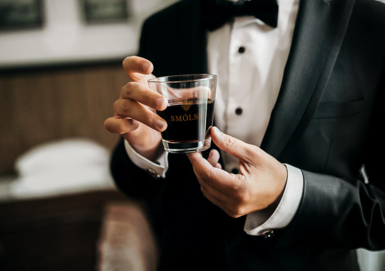 Mężczyzna w smokingu trzyma szklankę z napisem 'Smóls' w kolorze złotym, element identyfikacji wizualnej.