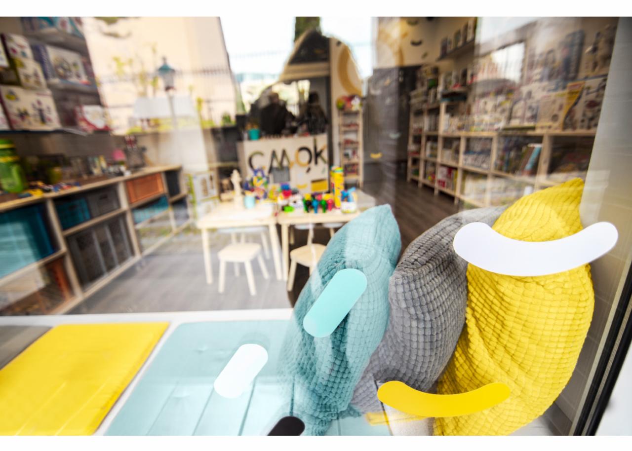 Witryna sklepu z zabawkami i warsztatowni w Poznaniu, widoczna przez szybę z naklejonymi elementami graficznymi w kształcie uśmiechów, wewnątrz regały z zabawkami i stoliki do zajęć.