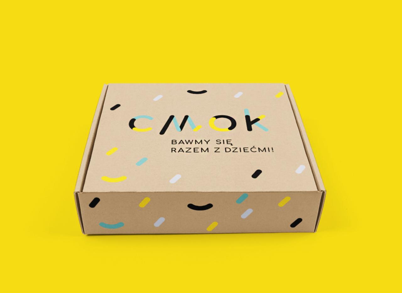 Kartonowe pudełko z logo 'CMOK' i napisem 'BAWMY SIĘ RAZEM Z DZIEĆMI!' na jaskrawym żółtym tle.