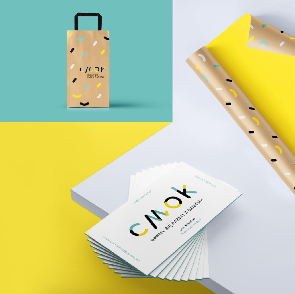 Projekt wizualny sklepu z zabawkami: torba papierowa z uchwytami, rolka papieru do pakowania i wizytówki z logo 'CMOK' na białym tle z elementami turkusowego i żółtego.