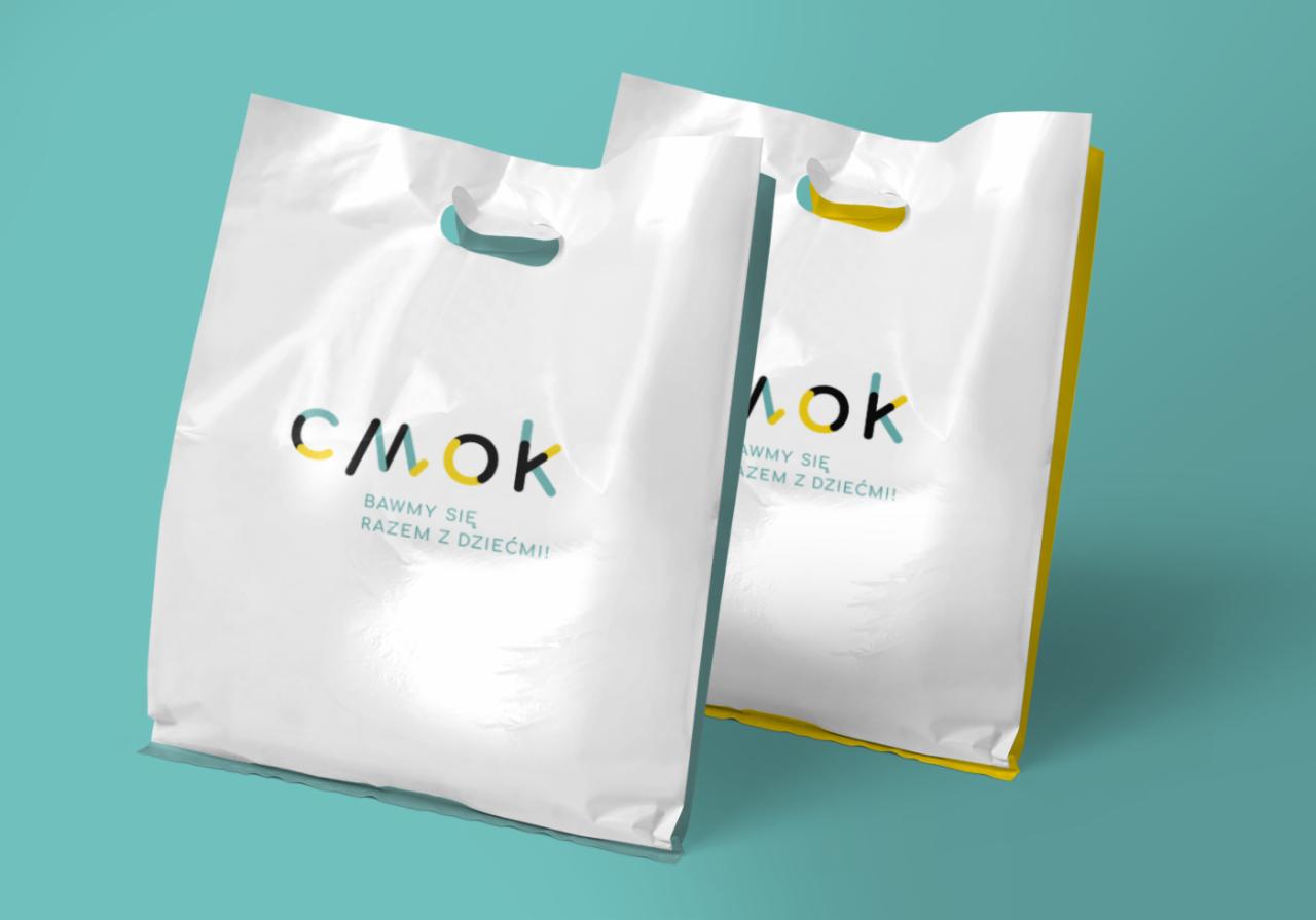 Dwie białe plastikowe torby z nadrukowanym logo 'SMOK' w kolorach czarnym, żółtym i turkusowym oraz hasłem 'BAWMY SIĘ RAZEM Z DZIEĆMI!', na turkusowym tle.