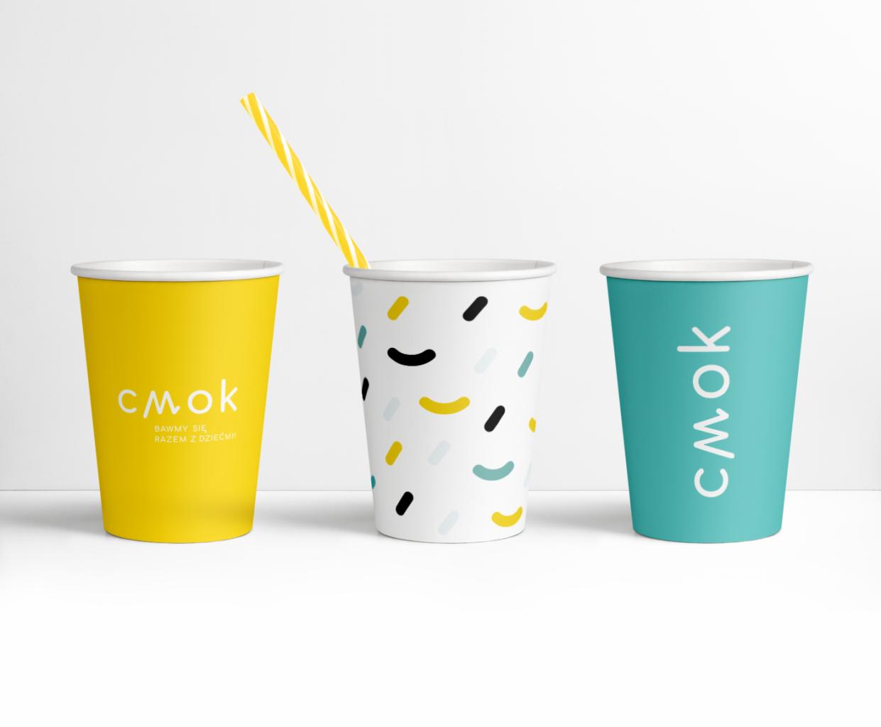 Trzy papierowe kubki z logo 'Smok' w kolorach żółtym, białym z kolorowymi wzorami i turkusowym, ustawione na białym tle, z żółtą słomką w środkowym kubku.