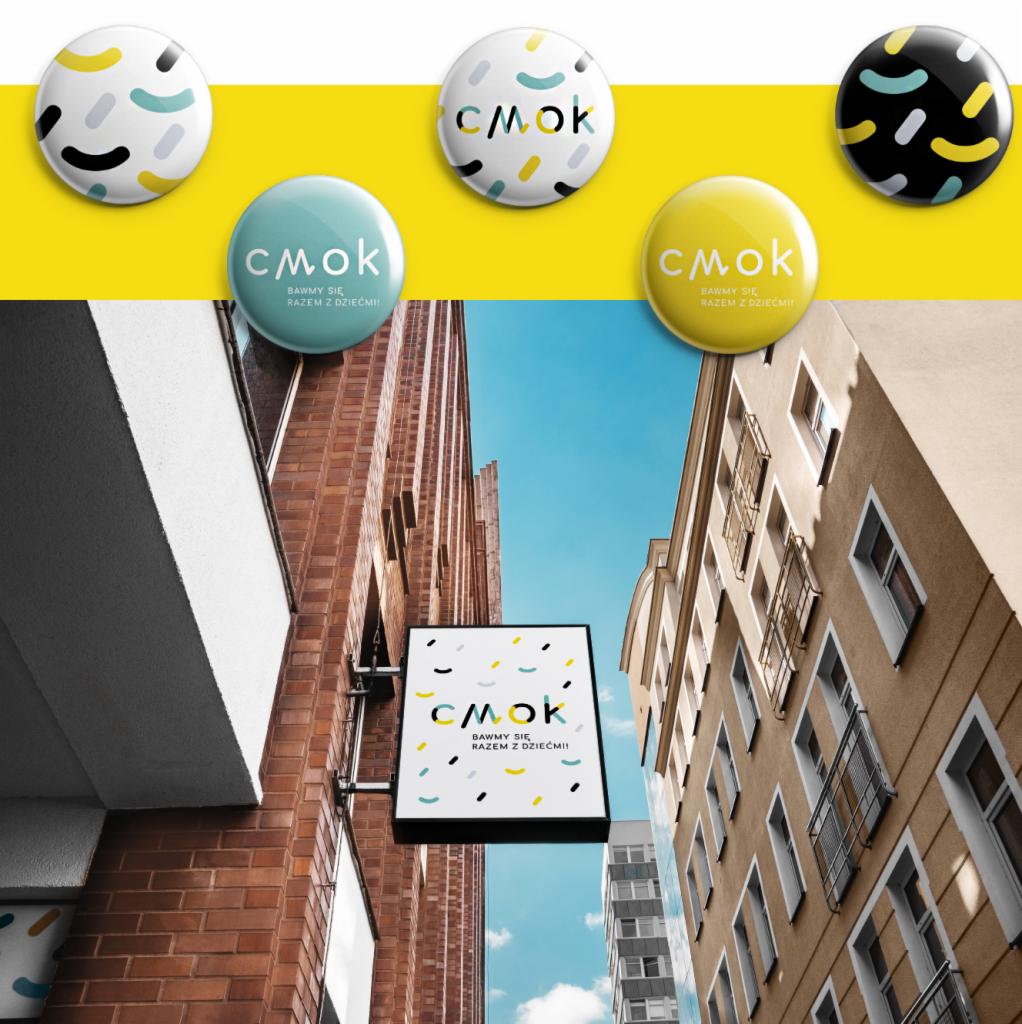 Tablica reklamowa z logo 'CMOK Bawmy się razem z dziećmi' umieszczona na elewacji budynku, z widocznymi budynkami w tle i zestawem okrągłych przypinek z wariantami tego samego logo na żółtym tle.