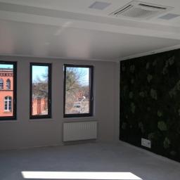 Maksimus - Wnętrze pokoju po pracach wykończeniowych, z trzema oknami z widokiem na ceglany budynek, panelem z mchu na ścianie i klimatyzacją sufitową.