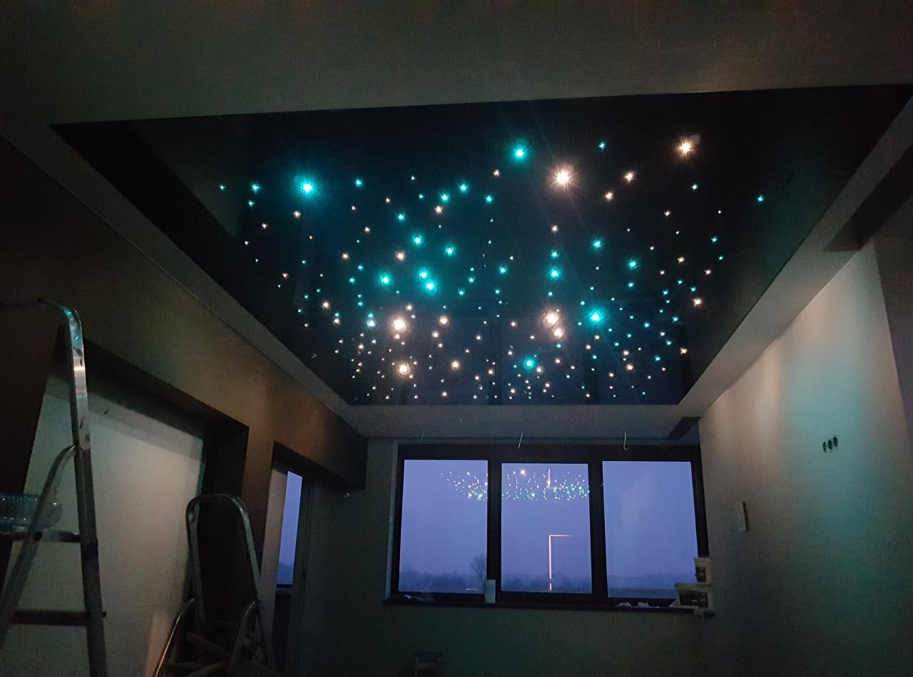 Czarny sufit podwieszany z oświetleniem LED imitującym gwiazdy, widok z dołu, drabina oparta o ścianę po lewej stronie, okno w tle.