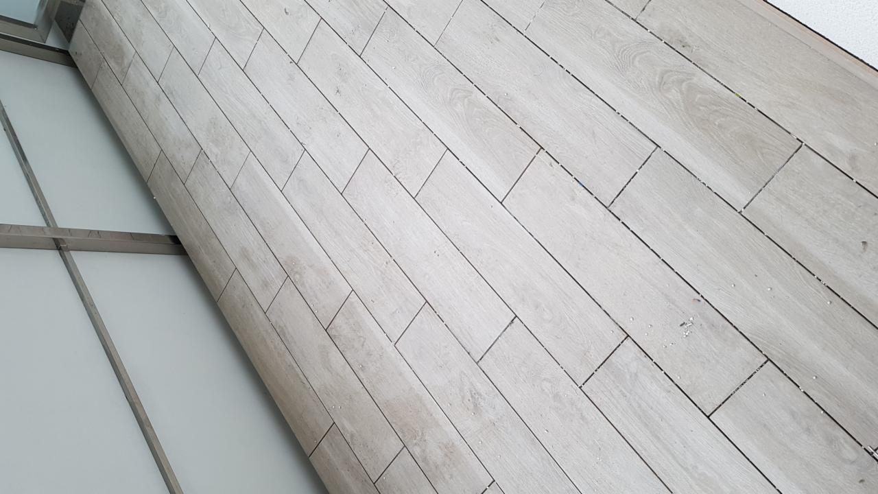 Ukośnie ułożone, jasne panele podłogowe imitujące drewno, widoczne drobne zabrudzenia w szczelinach, fragment okna z metalową ramą w lewym dolnym rogu.