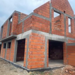 Smeton Sp. z o.o. - Surowy stan domu z czerwonej cegły w Otwocku, z widocznymi otworami okiennymi i betonowymi nadprożami, na tle pochmurnego nieba. Widoczny fundament i teren budowy.