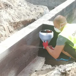 Pracownik w masce i kamizelce ochronnej, w czerwonych rękawicach, natryskowo zabezpiecza fundamenty przed wilgocią, widoczny sprzęt natryskowy z niebieskim przewodem.