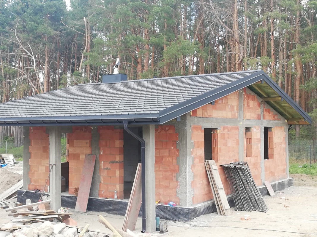 Niedokończony budynek z czerwonej cegły z szarym dachem w trakcie budowy, widoczne fundamenty, ściany i dach, na tle lasu.