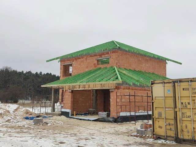 Dwukondygnacyjny dom murowany w trakcie budowy, z widoczną konstrukcją dachu z zielonych łat, ściany z czerwonej cegły, na zasnieżonym placu budowy, kontener magazynowy po prawej stronie.